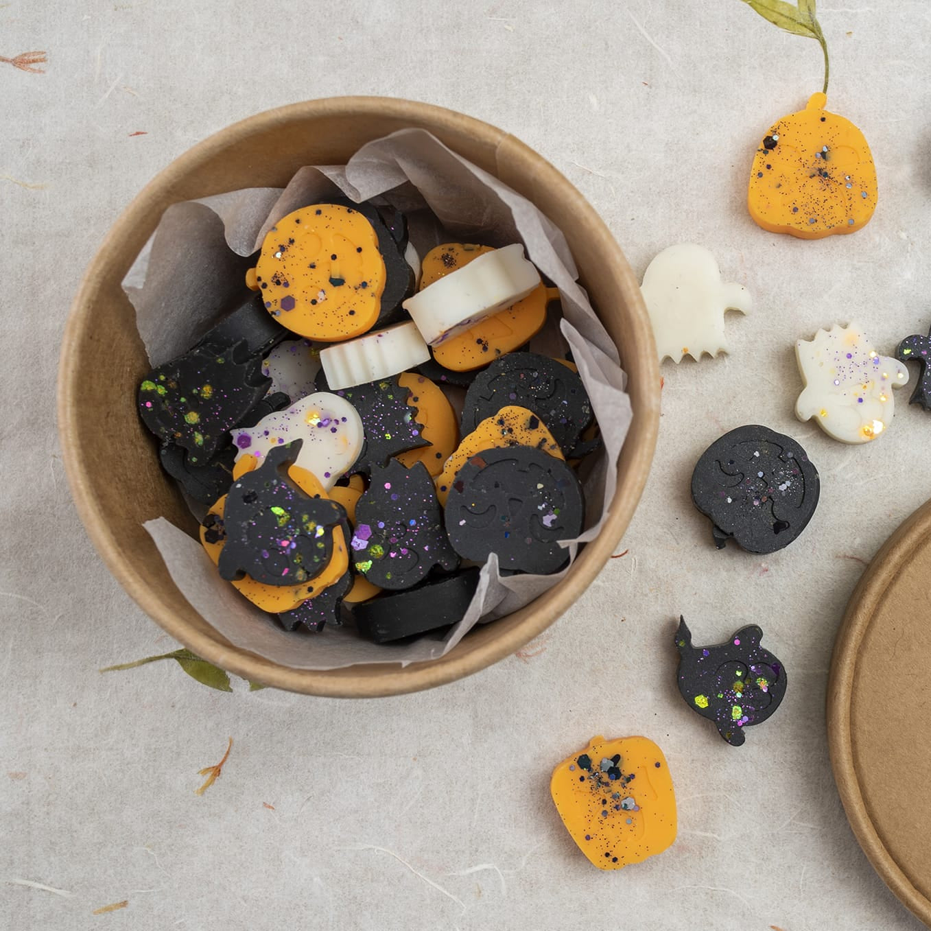 Una caja redonda de cartón de Fleur-de-Bois contiene Wax Melts Esencias en Miniatura CREEPY BOO PEAKY SPOOKY, con forma de calabazas, fantasmas y gatos en negro, naranja y blanco -algunos con purpurina- etiquetados como "Fleurs-de-Bois".