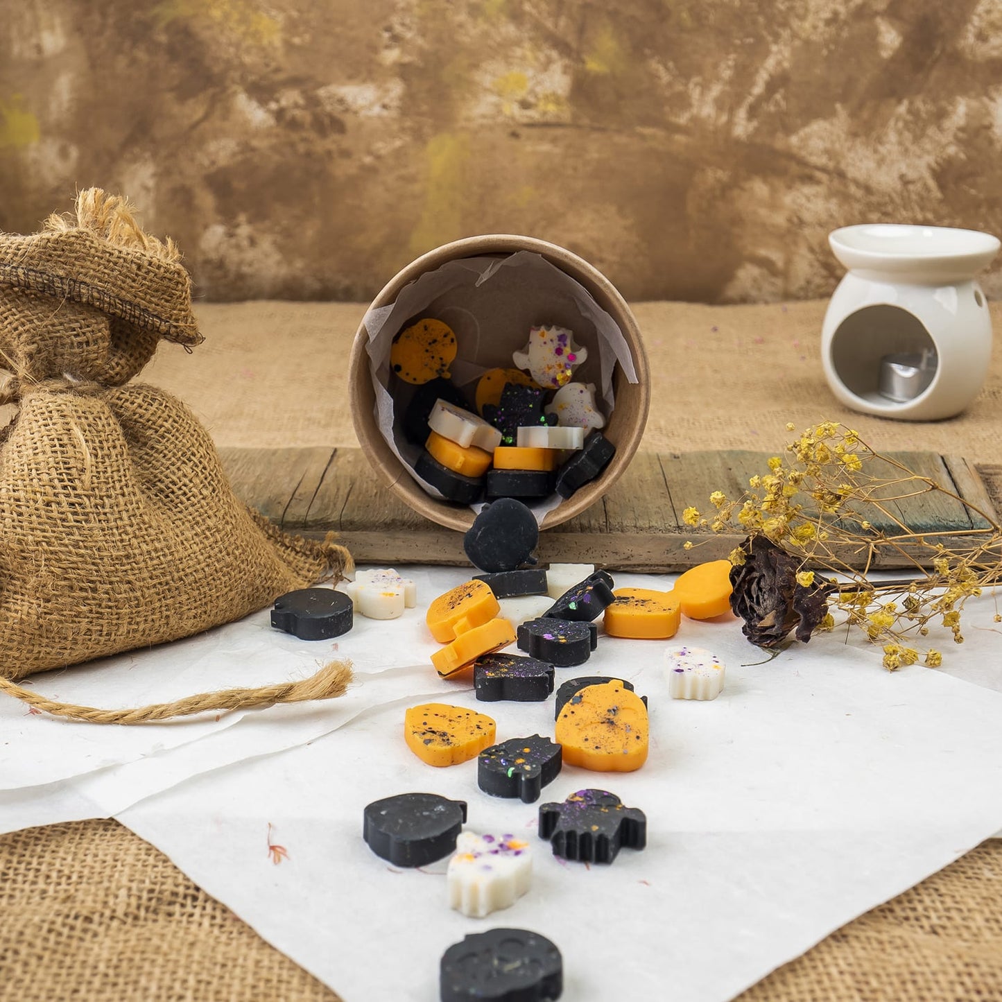 Una escena rústica con Wax Melts Esencias en Miniatura CREEPY BOO de Fleur-de-Bois, wax melts derramándose de un vaso de papel sobre papel blanco, junto a una bolsa de arpillera, flores secas y un quemador de aceite de cerámica sobre madera ante un fondo de textura marrón.
