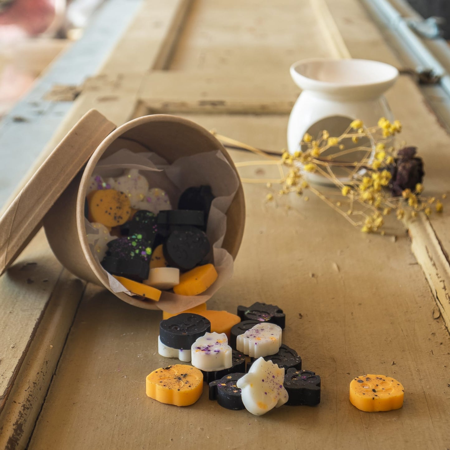 Un vaso de carton volcado con Fleur-de-Bois Wax Melts Esencias en Miniatura CREEPY BOO PEAKY SPOOKY-fantasmas y calabazas negras, blancas y naranjas que se derrama sobre madera rústica cerca de un calentador de cerámica blanca y flores secas.