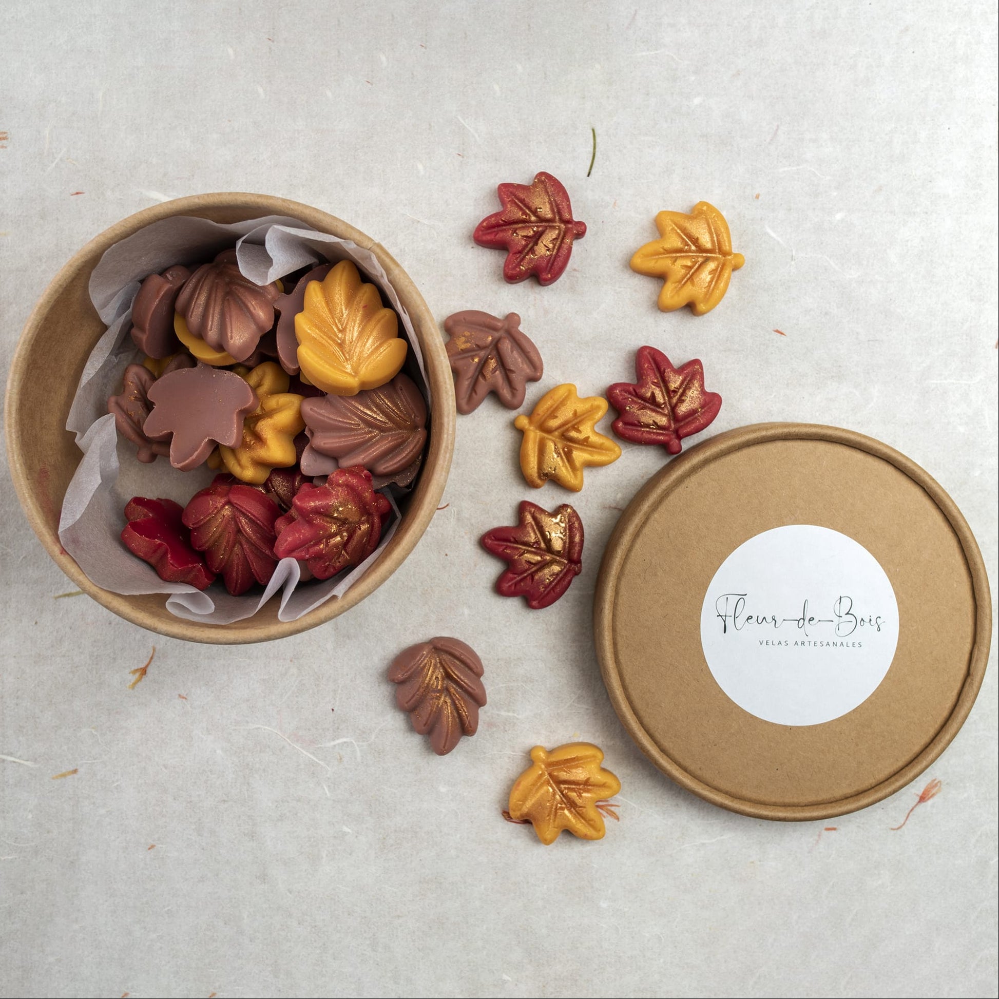 Una caja redonda de Melts de Cera Flor de Bois Esencias en Miniatura DELIRIOUS AUTUMN presenta melts de cera de soja con forma de hojas otoñales en marrón, rojo y amarillo. Algunas se esparcen por los alrededores, llenando el aire de evocadores aromas otoñales.