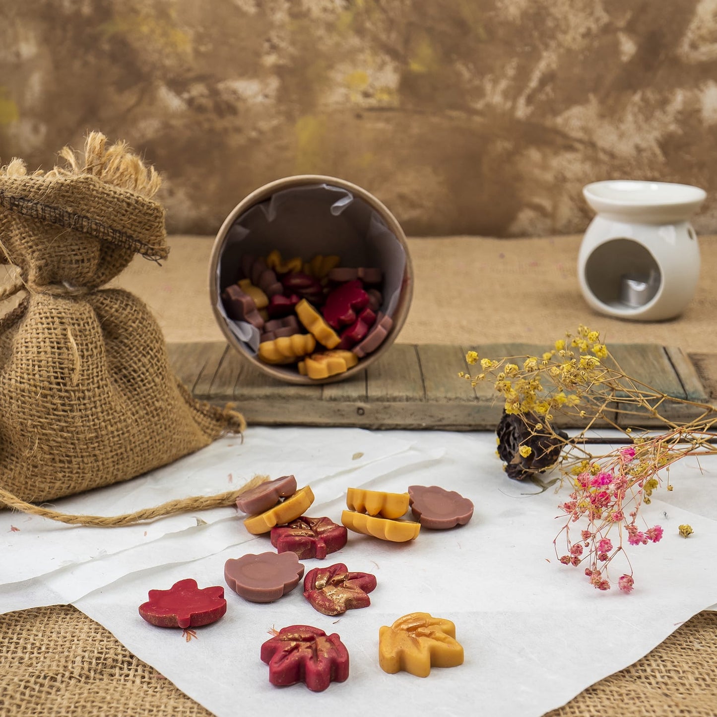 Una escena rústica presenta coloridos Wax Melts Esencias en Miniatura DELIRIOUS AUTUMN de Fleur-de-Bois, melts en forma de flor derramados de un recipiente, con flores secas, un saco de arpillera y un quemador de cera de cerámica blanca sobre madera.