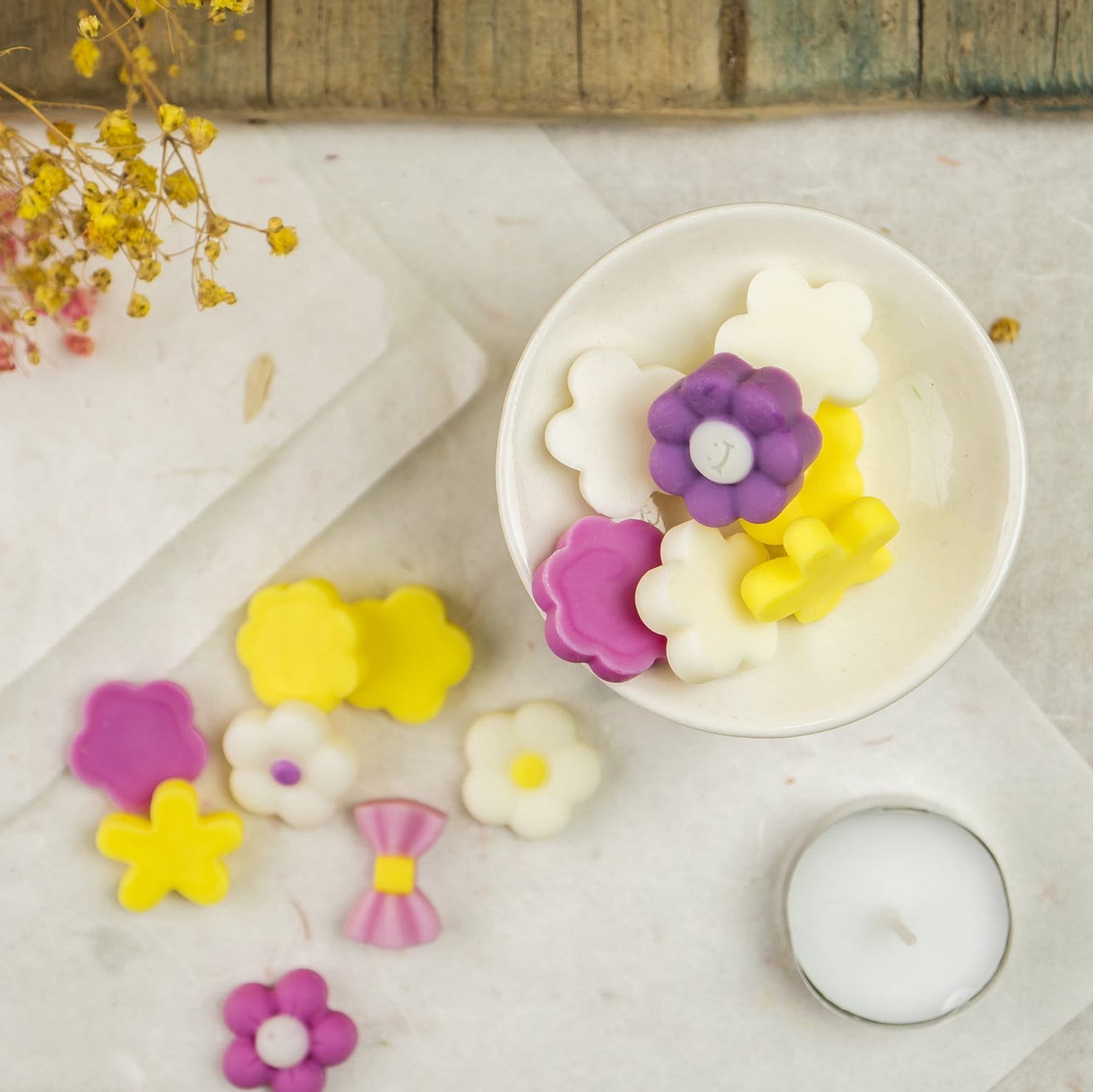 Una taza contiene coloridas Melts de Cera Esencias en Miniatura FLORES DE PRIMAVERA de Fleur-de-Bois en morado, amarillo y blanco, rodeadas de más melts en forma de flor inspirados en aromas primaverales, una candelita y flores secas sobre una superficie blanca.