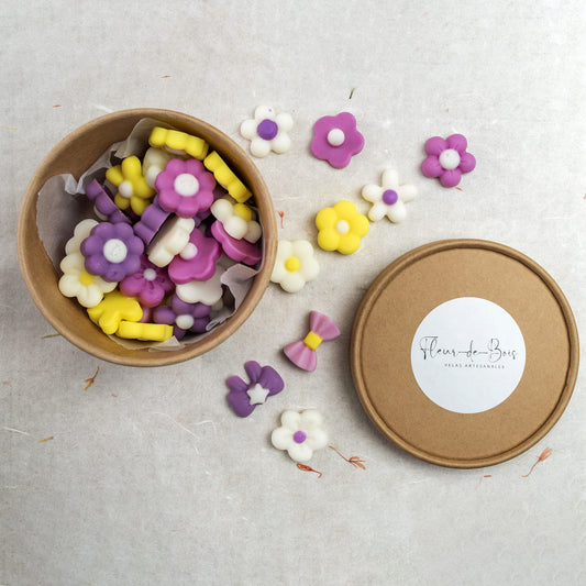 Una caja redonda de Esencias en Miniatura FLORES DE PRIMAVERA de Fleur-de-Bois contiene esencias de soja en forma de flor en amarillo, morado y blanco. En el exterior hay fundentes adicionales y un lazo morado, y la tapa "Fleur-de-Bois" descansa a la derecha.