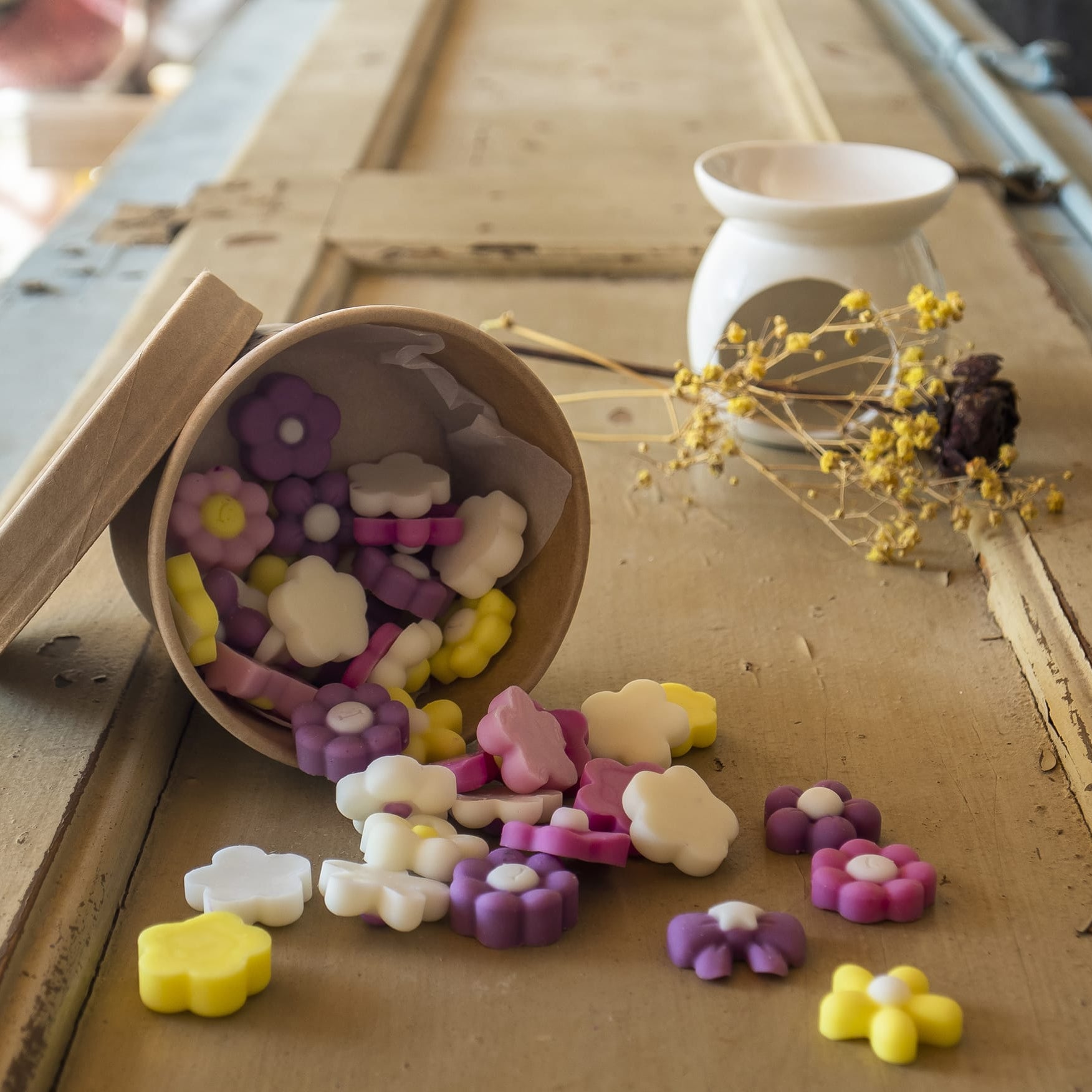 Coloridas Esencias en Miniatura FLORES DE PRIMAVERA de Fleur-de-Bois se derraman desde una caja de cartón sobre madera, con un calentador de cera blanco y flores secas de fondo que evocan fragancias otoñales.