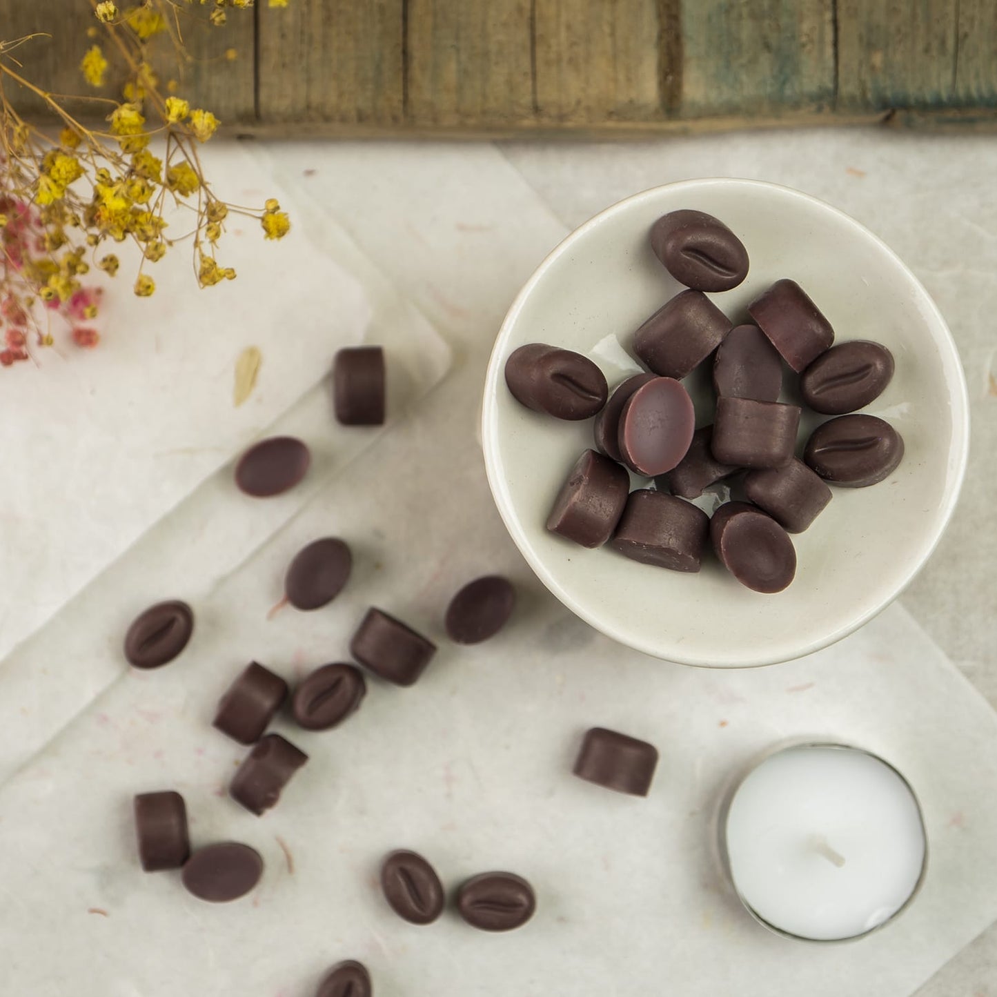 Un cuenco blanco contiene Melts de Cera Flor de Bois Esencias en Miniatura GRANOS DE CAFÉ DOLCE CAFFÉ, algunos con forma de granos de café, sobre una superficie clara con melts dispersos cerca. Una vela de cera de soja y flores amarillas secas completan la escena.