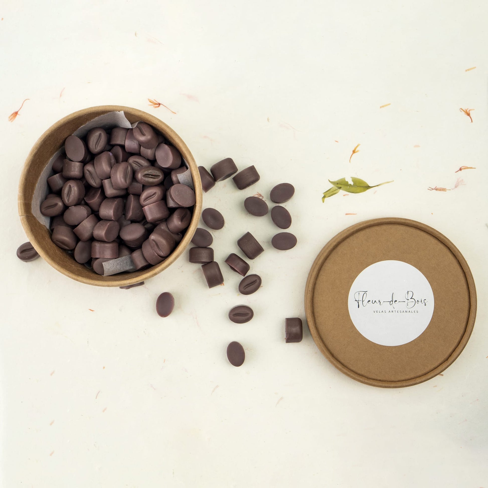 Una caja redonda de papel kraft abierta con la etiqueta "Fleur-de-Bois" revela Melts de Cera Esencias en Miniatura GRANOS DE CAFÉ DOLCE CAFFÉ, algunos con forma de aromáticos granos de café, con melts esparcidos sobre una superficie blanca junto a la tapa.