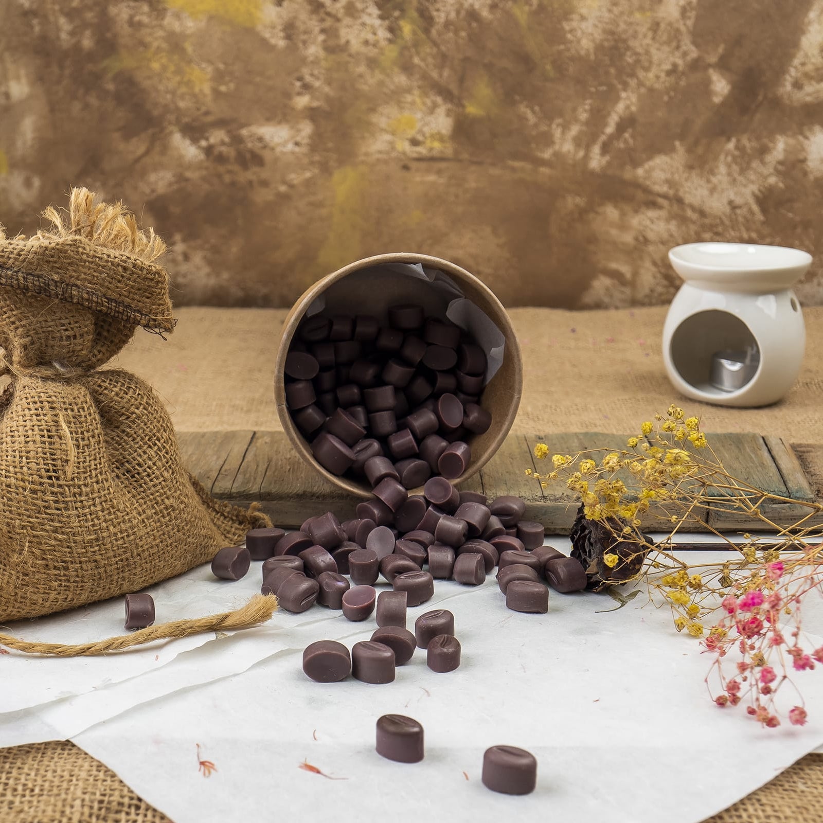 Cera Fundida Esencias en Miniatura GRANOS DE CAFÉ DOLCE CAFFÉ de Fleur-de-Bois se derraman sobre una superficie blanca junto a un saco de arpillera, aromáticos granos de café, flores secas y un portavelas de cerámica blanca sobre un fondo rústico.
