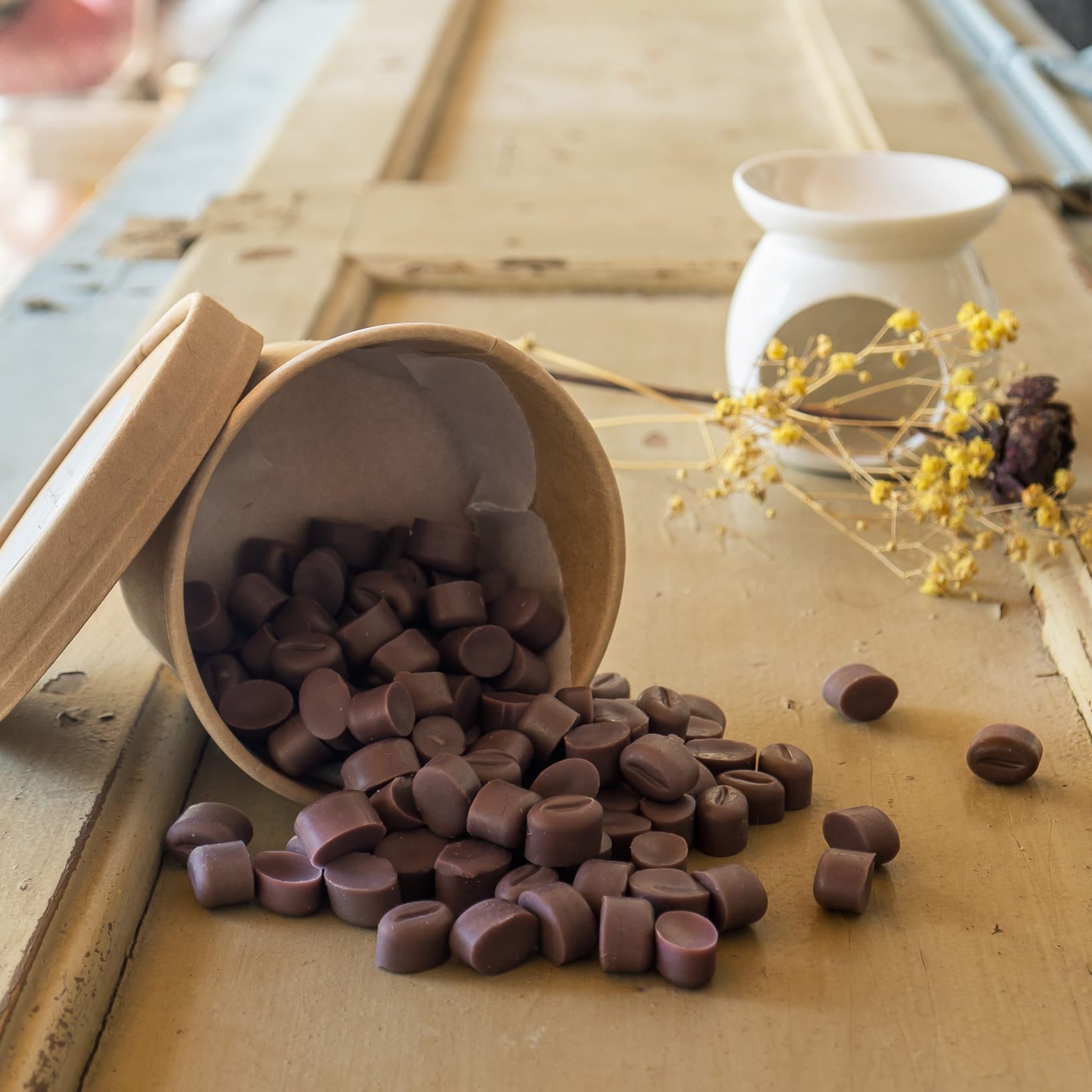 Un recipiente marrón de Flor de Bois Wax Melts Esencias en Miniatura GRANOS DE CAFÉ DOLCE CAFFÉ se derrama sobre una superficie de madera, con flores amarillas secas y una taza de cerámica blanca al fondo.