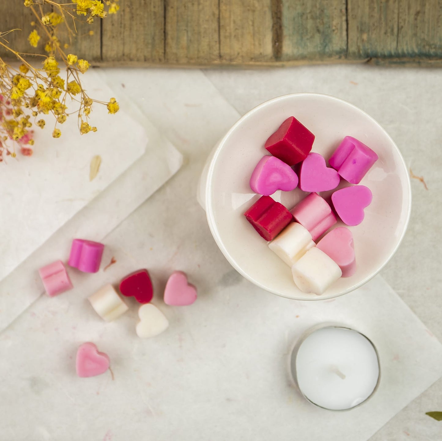 Un cuenco blanco contiene Melts de Cera Esencias en Miniatura Corazón Deseos en rosa, rojo y blanco, en forma de corazón y de cubo, sobre mármol junto a una candelita y melts esparcidos. Flores secas amarillas asoman por una esquina.