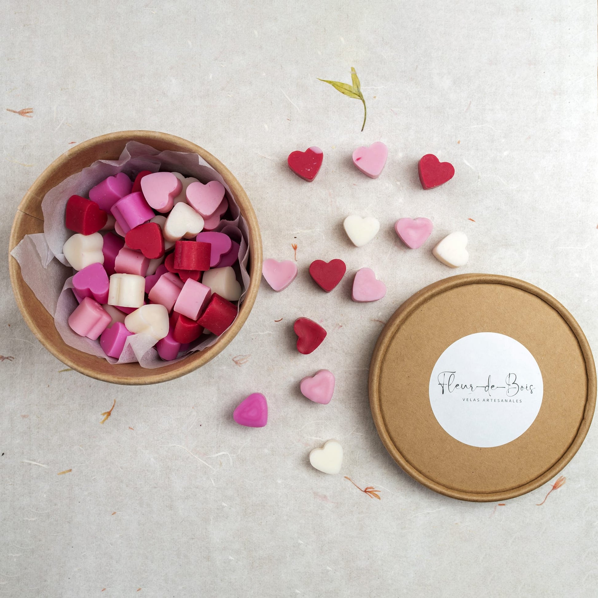 Una caja redonda y marrón de Esencias en Miniatura CORAZÓN DESEOS, rellena de cera fundida de soja (cera de soja) rosa, roja y blanca en forma de corazón, descansa sobre una superficie clara. La tapa descansa cerca, evocando aromas de flores de primavera.