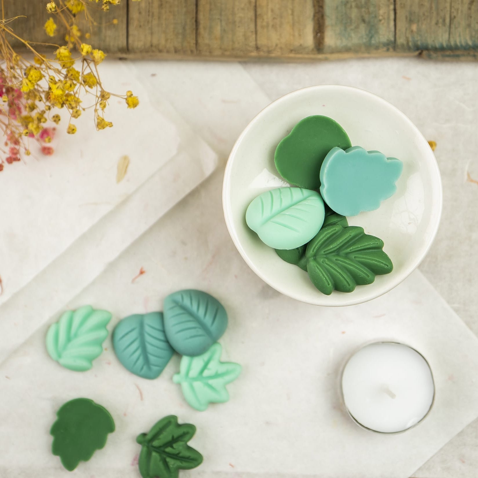 Un quemador blanco contiene wax melts Fleur-de-Bois Esencias en Miniatura HOJAS SUELTAS, con forma de hoja en cera de soja verde y turquesa. Más ceras fundidas, una vela candelita y flores secas decoran la rústica superficie de madera cercana.