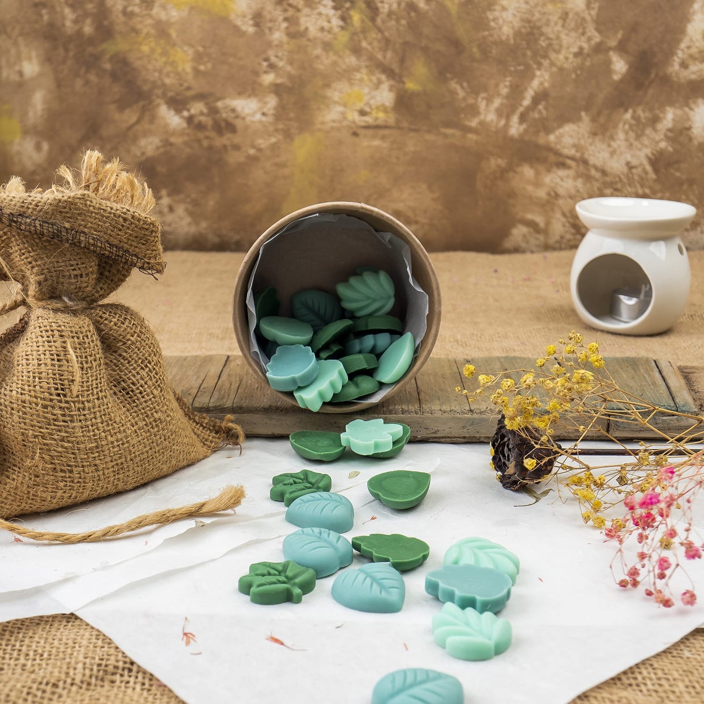 Una pequeña bolsa de tela y un cubo con punta derraman wax melts verde y azul Esencias en Miniatura HOJAS SUELTAS Fleur-de-Bois sobre una tabla de madera, con flores secas y un calentador de cerámica blanca al fondo.