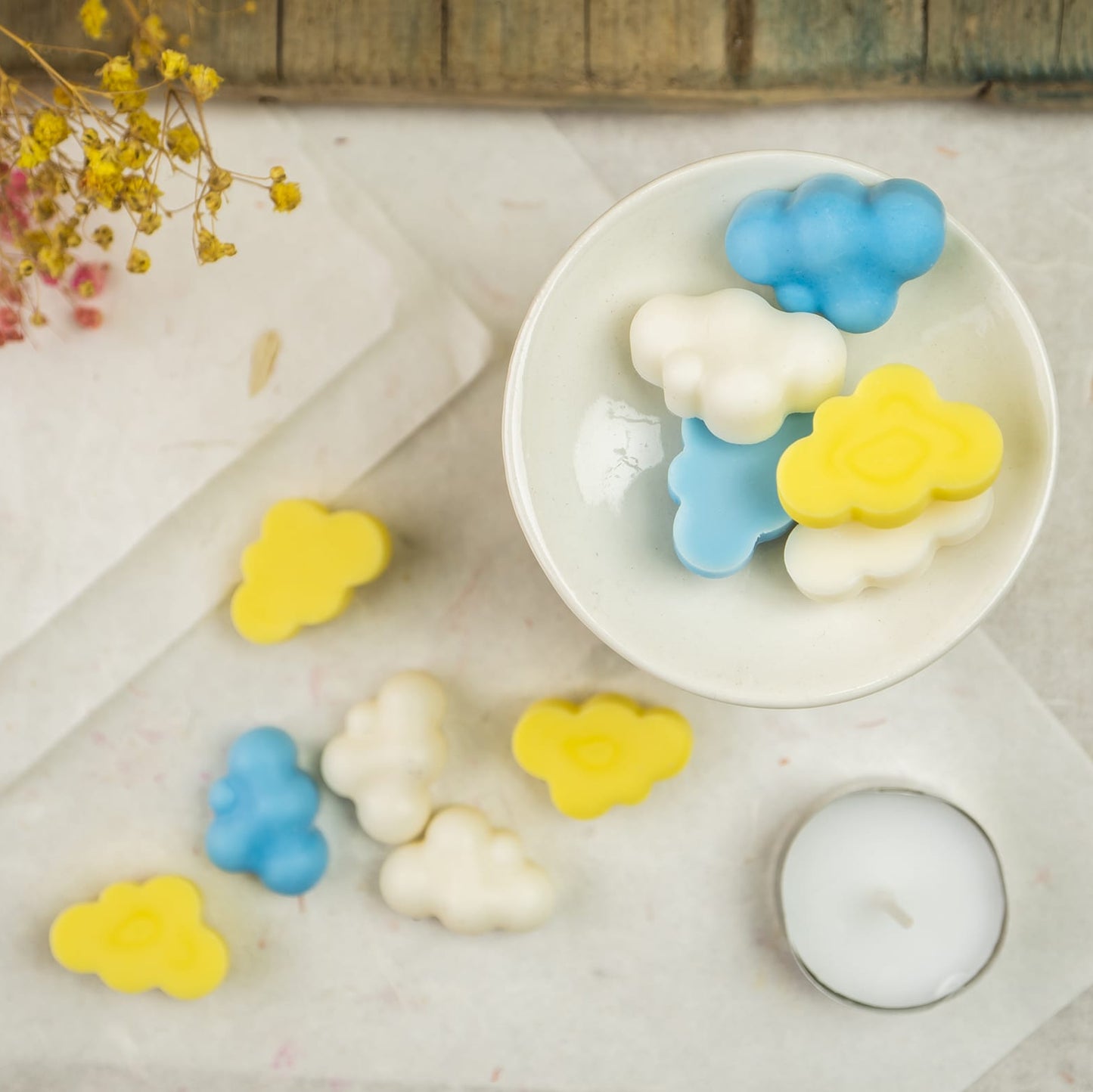 Un quemador de cera blanco con Fleur-de-Bois Esencias en Miniatura NUBES DE ALGODÓN en azul, amarillo y blanco está sobre una mesa con hojas de esencias, una vela candelita, fragancias naturales y flores secas.