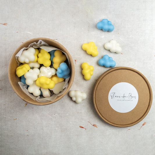 Una caja redonda de color marrón etiquetada como Fleur-de-Bois contiene Wax Melts Esencias en Miniatura NUBES DE ALGODÓN de color blanco, amarillo y azul. Cerca, sobre una superficie clara, hay esparcidas varias ceras fundidas de soja.