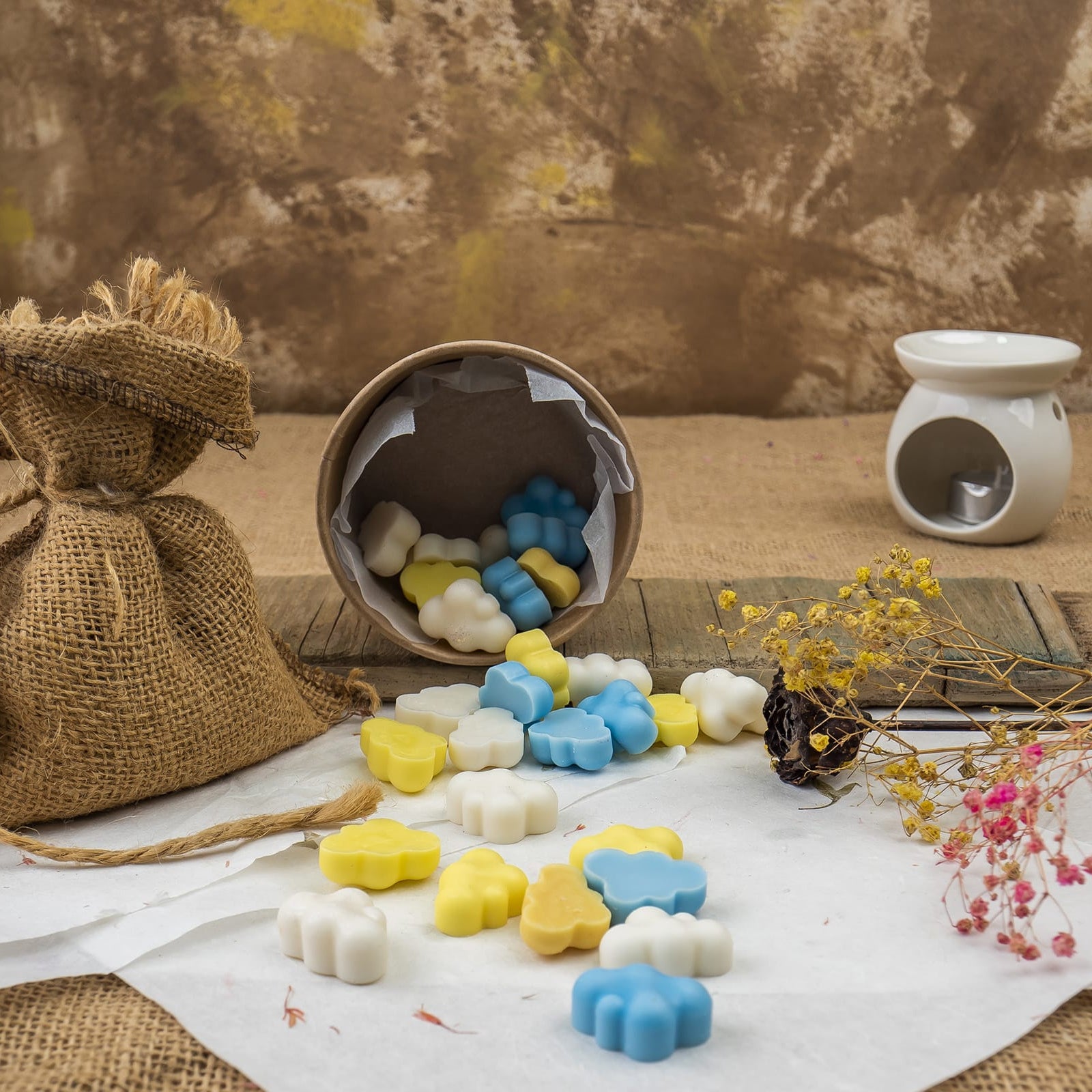 Un vaso con punta derrama fusiones de cera azules, amarillas y blancas Esencias en Miniatura NUBES DE ALGODÓN Fleur-de-Bois de Fleur-de-Bois, junto a flores secas y un quemador de aceite blanco sobre marrón rústico evocan fragancias naturales.