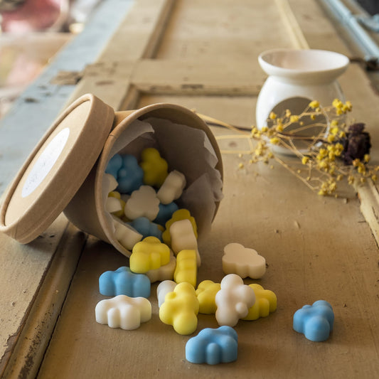 Un pequeño recipiente derrama Wax Melts azul, amarillo y blanco Esencias en Miniatura NUBES DE ALGODÓN Fleur-de-Bois sobre una superficie de madera rústica, con un calentador de cera blanco y flores secas de Fleur-de-Bois de fondo.